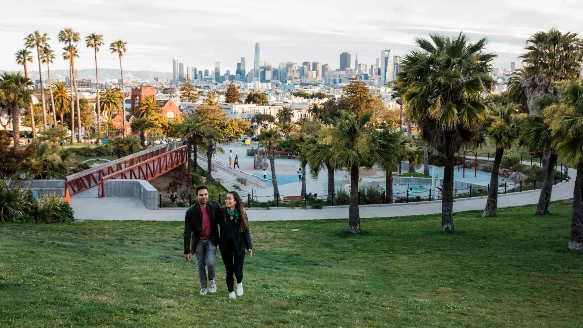 Um casal caminha em direção à câmera com Dolores Park e o horizonte de São Francisco atrás deles.